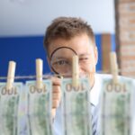 Pranie pieniędzy. Mężczyzna patrzy przez lupę na sto dolarowe wiszące na spinaczach do bielizny.