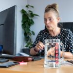 Kobieta w średnim wieku pracująca w biurze na stole na pierwszym planie szklany kubek z czystą wodą