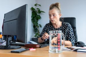 Kobieta w średnim wieku pracująca w biurze na stole na pierwszym planie szklany kubek z czystą wodą
