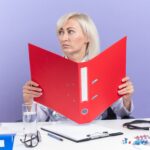 Pewna dorosła słowiańska lekarka w szacie medycznej ze stetoskopem siedząca przy biurku z narzędziami biurowymi trzymającymi folder plików i patrząca na bok na fioletowym tle z kopią miejsca