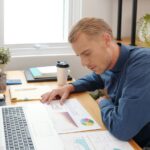 Zamyślony młody przedsiębiorca sprawdzający i analizujący informacje w dokumentach finansowych na swoim biurku