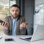 Biznesmen w nowoczesnym biurze wydaje się zdezorientowany, trzymając smartfon i wiele kredytów