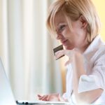 Młoda uśmiechnięta kobieta w domu używa komputera do zakupów internetowych, trzymając w ręku kartę kredytową