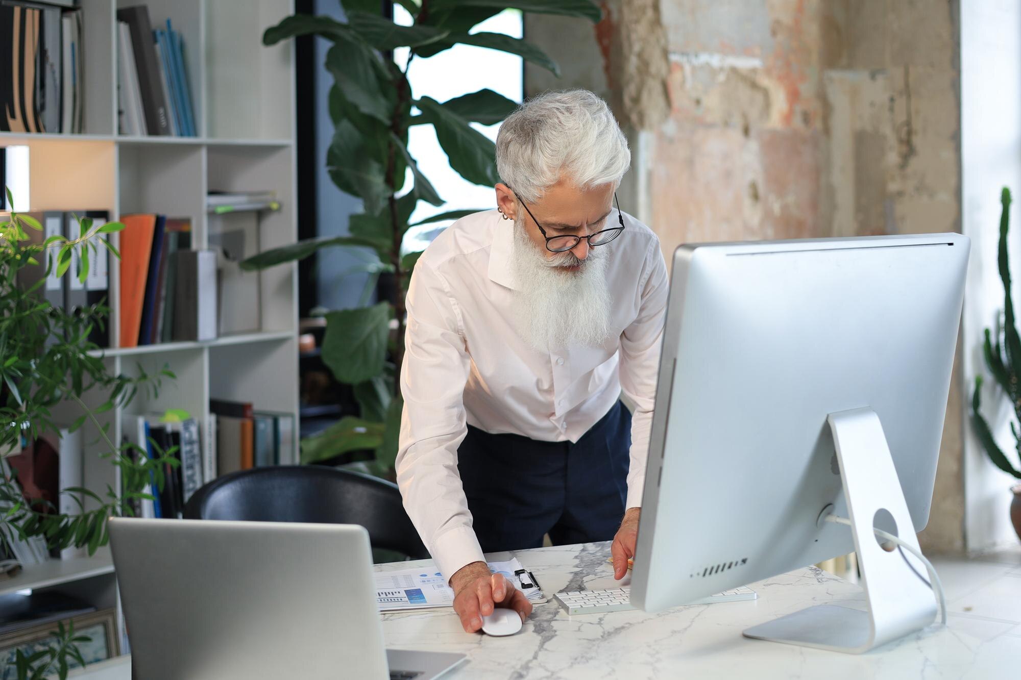 Starszy biznesmen ze stylową brodą pracuje na laptopie przy biurku w biurze