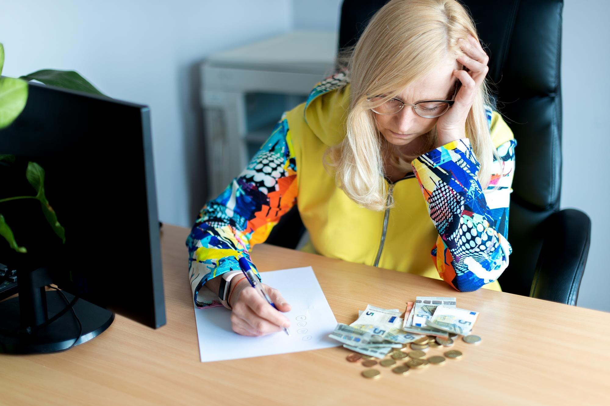 Zamyślona kobieta w średnim wieku licząca pieniądze w euro i robiąca notatki przy biurku w domowym biurze