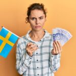 Młoda brunetka trzyma flagę szwecji i banknoty korony sceptyczna i nerwowa, marszcząc brwi zdenerwowana z powodu problemu. negatywna osoba.