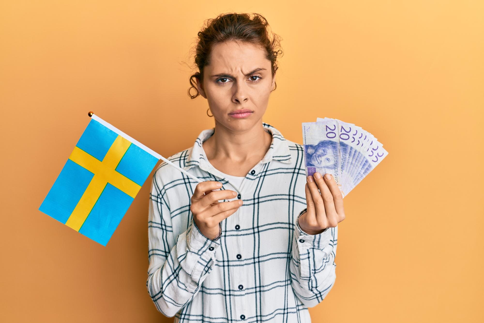 Młoda brunetka trzyma flagę szwecji i banknoty korony sceptyczna i nerwowa, marszcząc brwi zdenerwowana z powodu problemu. negatywna osoba.