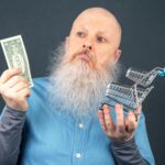 Mężczyzna z dużą białą brodą trzyma dolara i model kosza spożywczego w rękach.