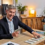 Happy young caucasian successful businessman in formal suit counting money cash at home office desk