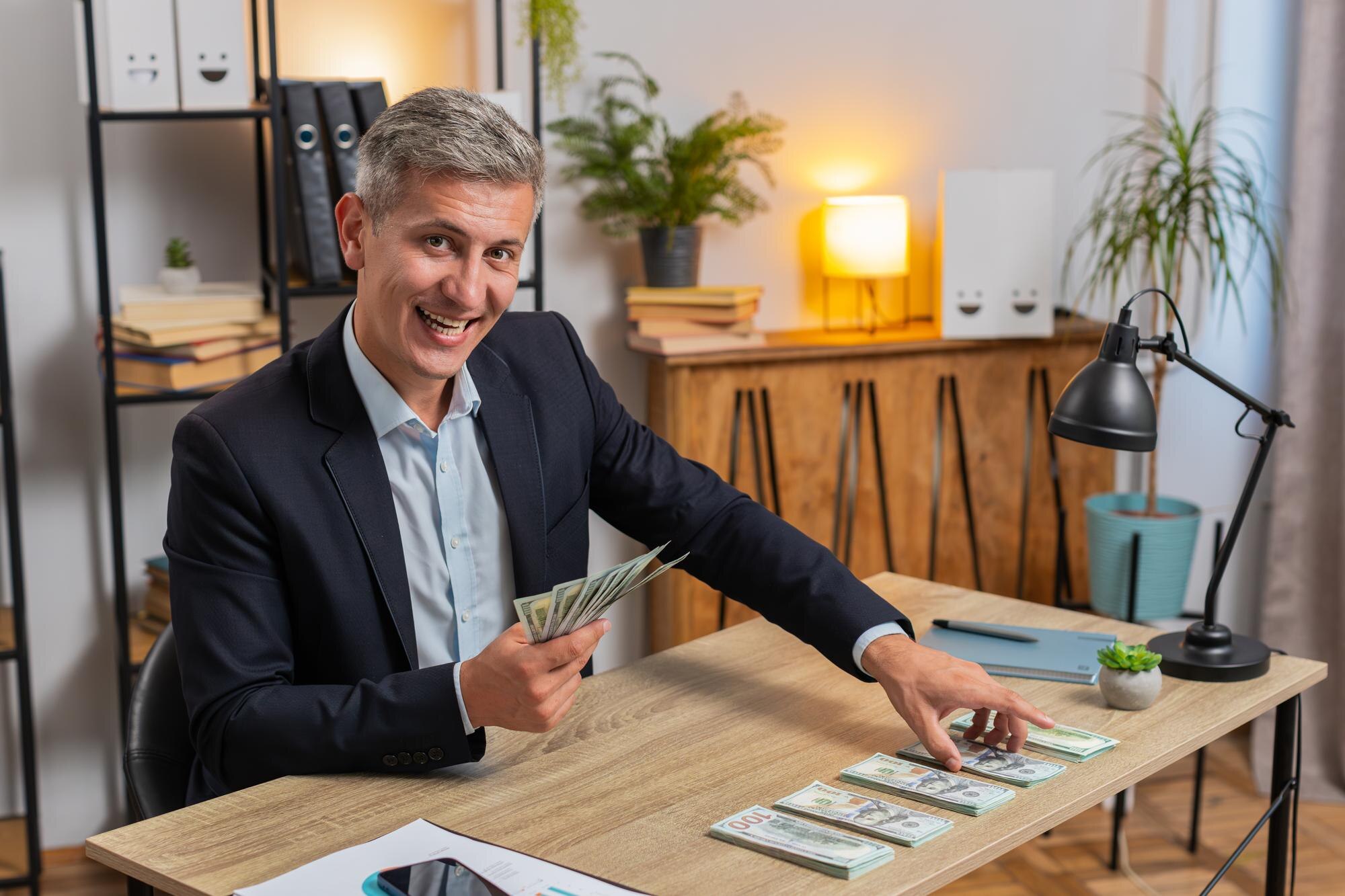 Happy young caucasian successful businessman in formal suit counting money cash at home office desk