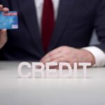 Mężczyzna w garniturze trzyma kartę kredytową przed białymi literami z napisem kredyt.