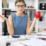 Młoda dziewczyna siedzi przy stole w biurze, trzymając w ręce czerwony kubek i pracując z dokumentami.